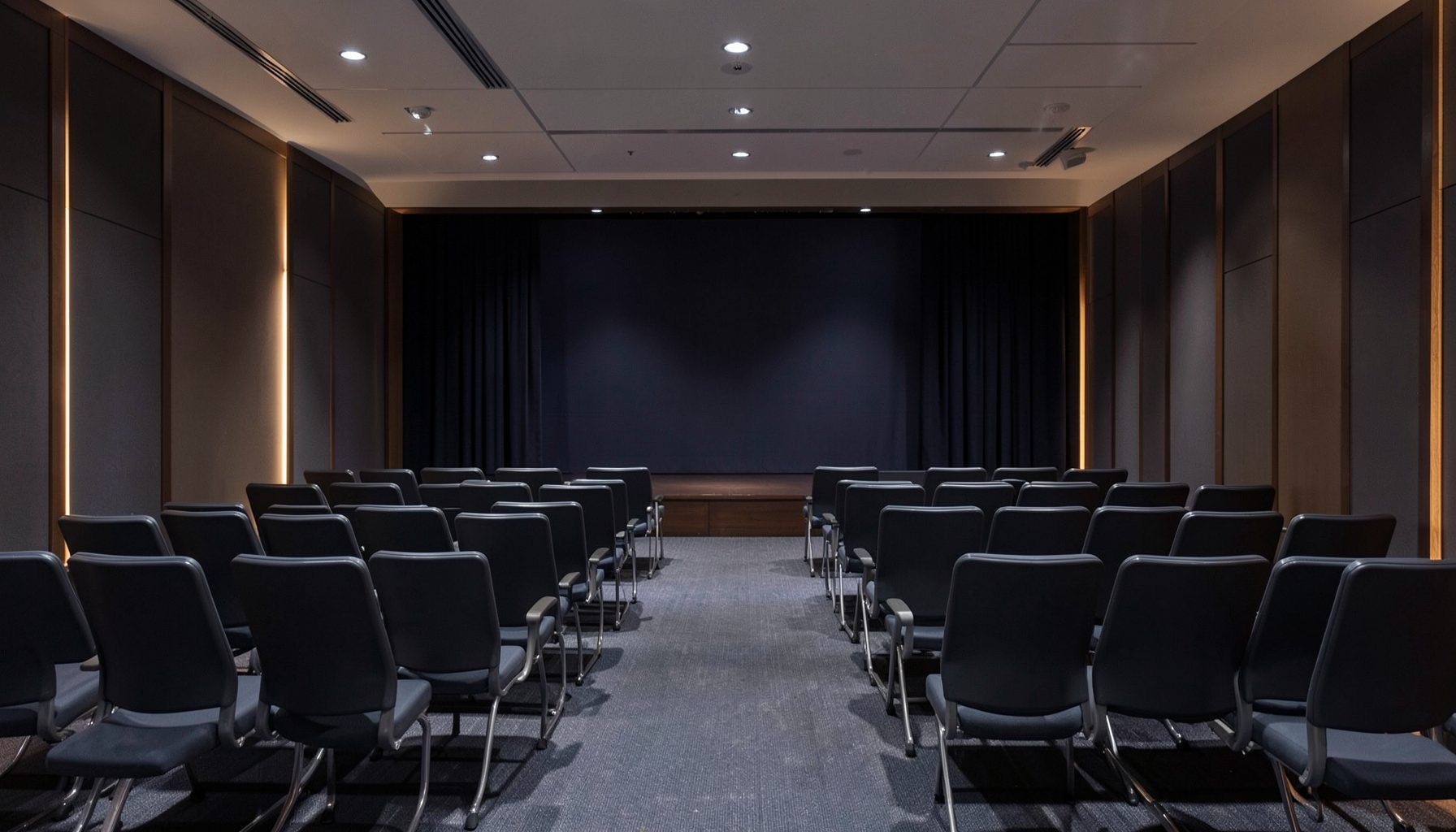 A softly lit conference room with rows of empty chairs and a stage in the background, deep navy and charcoal tones with warm amber accents