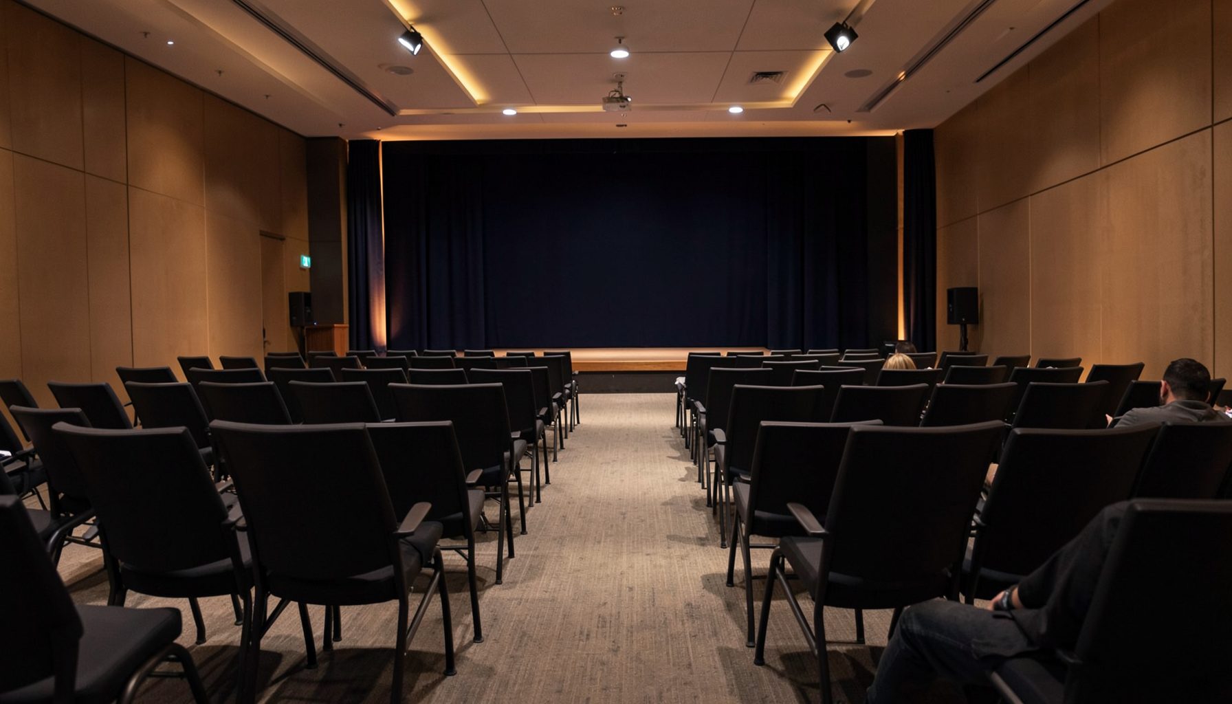 A softly lit conference hall with rows of empty chairs and a stage in the background, deep navy and warm amber tones blending in a quiet, professional atmosphere.