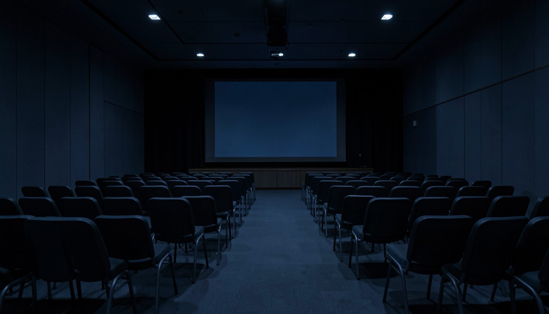 A dimly lit conference hall with rows of empty chairs facing a stage, deep navy and slate blue tones, quiet and anticipatory atmosphere